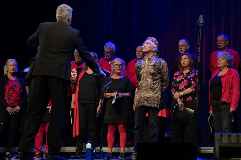 Een koor dat optreedt onder begeleiding van een dirigent, met verschillende zangers in kleurrijke kleding.