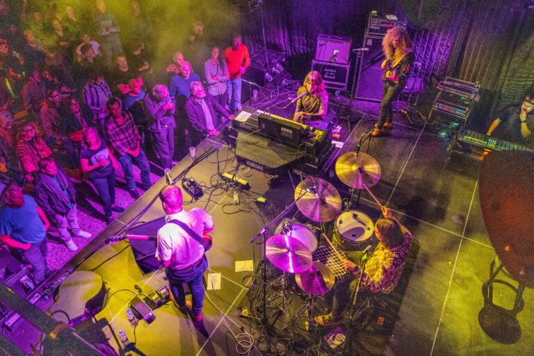 Een band speelt live op het podium voor een enthousiast publiek, omringd door kleurrijke verlichting.