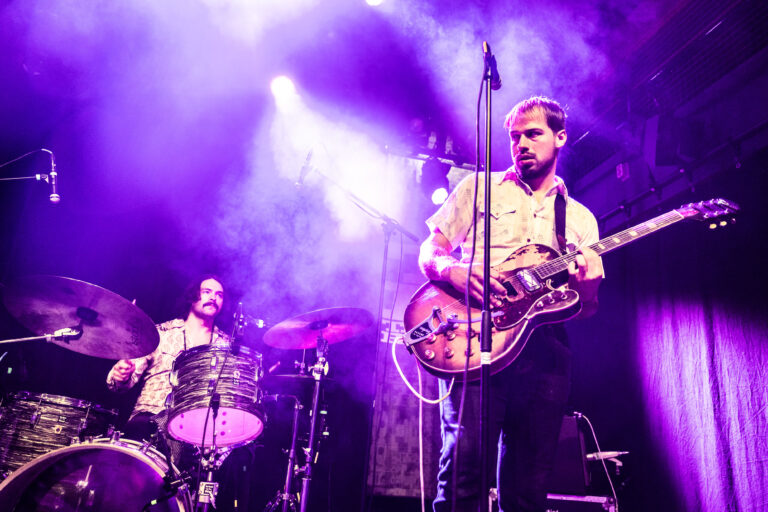 Twee muzikanten spelen op een podium met paarse verlichting, een van hen met een elektrische gitaar en de ander achter een drumstel.