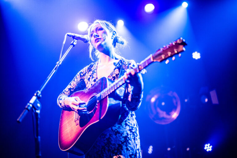 Een singer-songwriter performt op het podium met een akoestische gitaar, omringd door kleurrijke lichten.