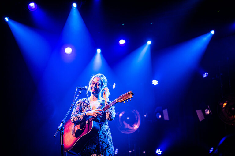 Zangeres met een gitaar staat op het podium onder blauwe lichten.