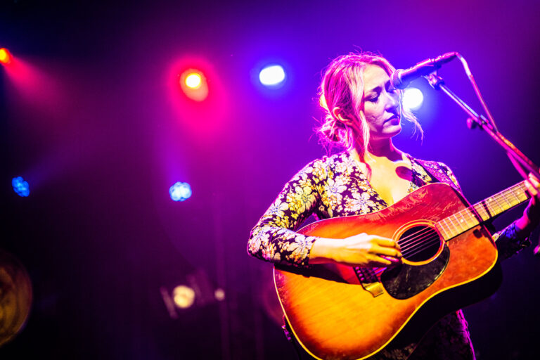 Een vrouwelijke muzikant speelt gitaar op het podium, omringd door kleurrijk licht.
