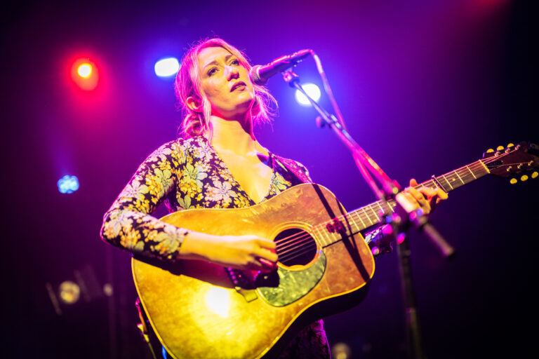 Een vrouw zingt en speelt gitaar op een podium, omringd door kleurrijke lichten.