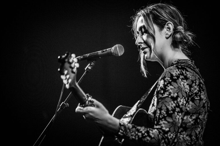 Een muzikante zingt en speelt gitaar op een podium met een microphone in beeld.