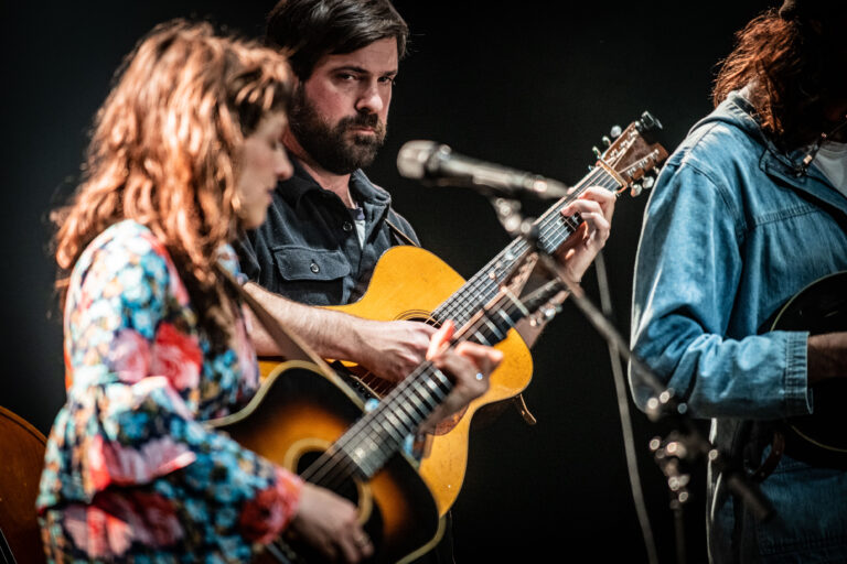 Een man en een vrouw spelen samen muziek op een podium met gitaren, terwijl ze naar elkaar kijken.