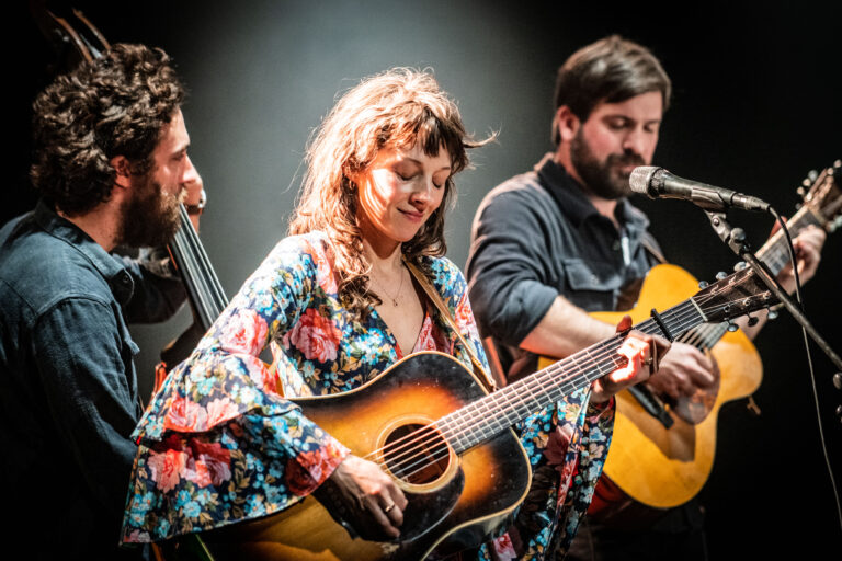 Drie muzikanten spelen samen op een podium, met een vrouwelijke zangeres in het midden die een gitaar vasthoudt.