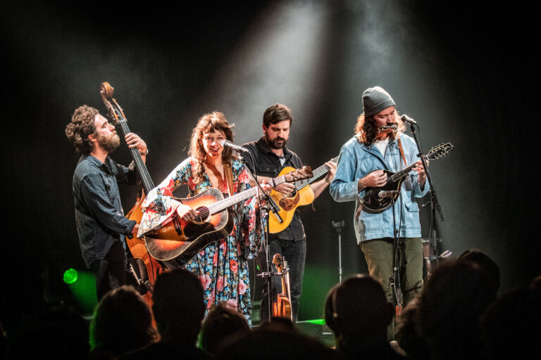 Een muziekband voert een optreden uit op het podium, omringd door lichtstralen en een publiek.