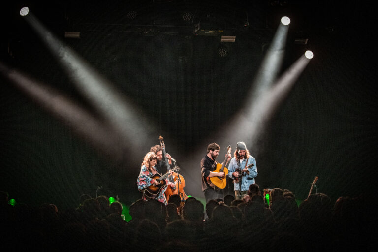 Een muziekgroep speelt op een podium met spotlights, terwijl het publiek eromheen kijkt.