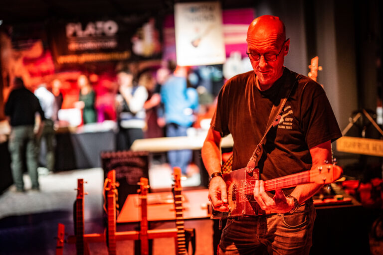 Een muzikant speelt een elektrische gitaar op een evenement, met mensen op de achtergrond.