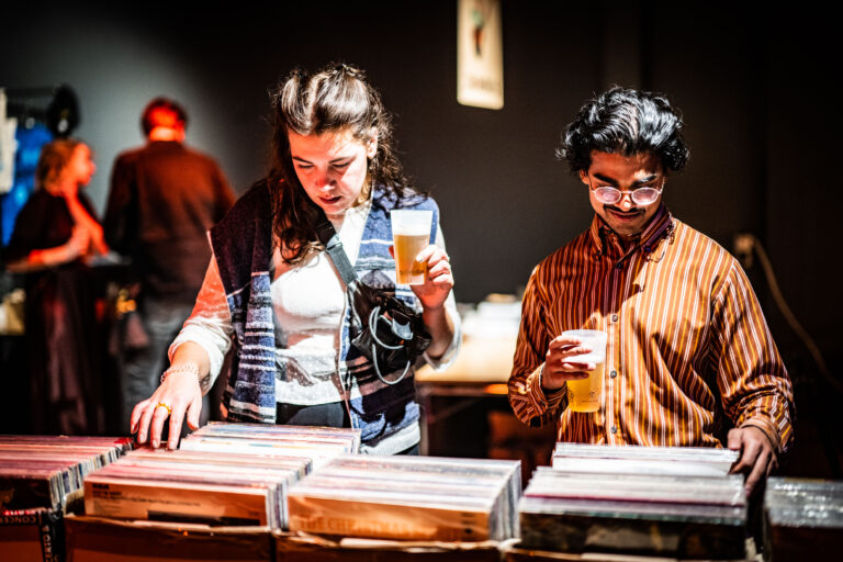 Twee mensen kijken naar vinylplaten terwijl ze een drankje vasthouden in een sfeervolle omgeving.
