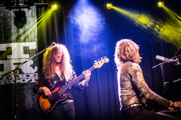 Een band speelt live op het podium met kleurrijke verlichting, waarbij een muzikant met een elektrische basgitaar in de schijnwerpers staat.