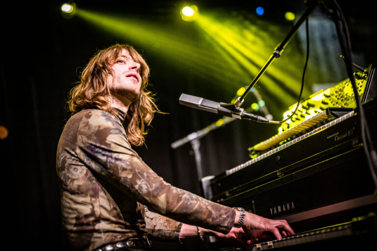 Een muzikant speelt op een piano terwijl ze naar het publiek kijkt, omringd door kleurrijke lichtstralen.