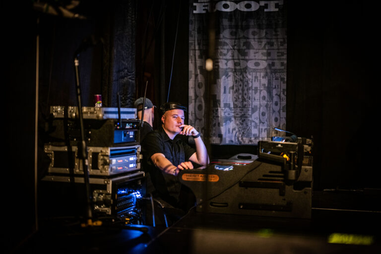 Een man zit achter een mengpaneel in een controlekamer, omringd door audioapparatuur en kabels.