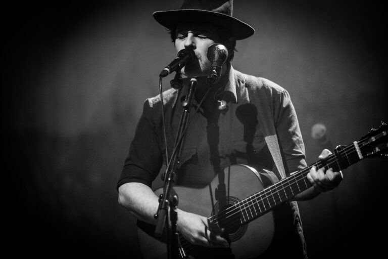 Zwart-wit foto van een muzikant die een akoestische gitaar speelt en zingt op een podium.