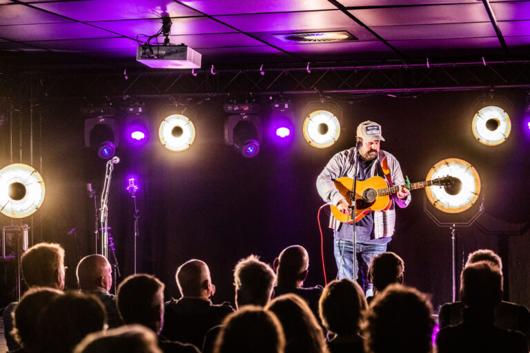 Een muzikant staat op een podium met een gitaar, terwijl een publiek hem aankijkt onder lichten in paarse tinten.