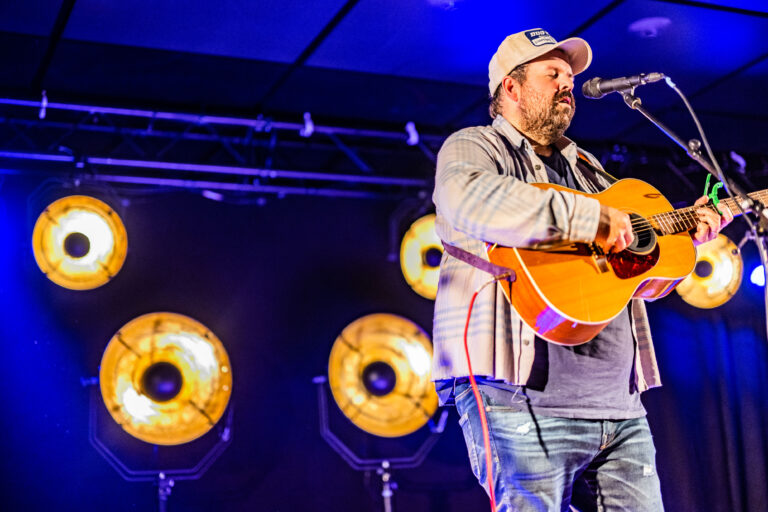 Een muzikant zingt en speelt gitaar op een podium, met opvallende lichten op de achtergrond.