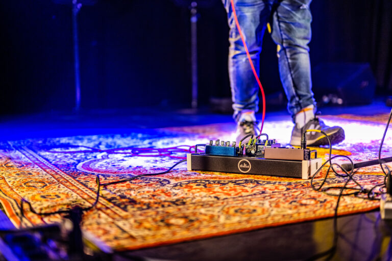 Een close-up van de voeten van een muzikant op een podium, met een effectpedalenset en kleurrijke verlichting.