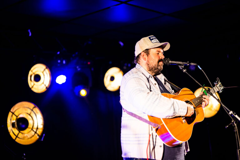 Een muzikant met een gitaar staat op het podium, omgeven door kleurrijke lichten.