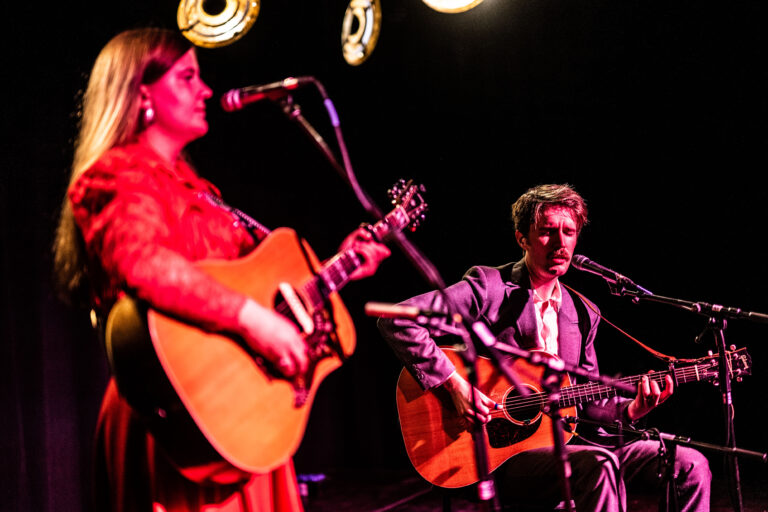 Twee muzikanten spelen op een podium, één met een gitaar en de ander met een elektrische gitaar, met sfeervolle verlichting op de achtergrond.