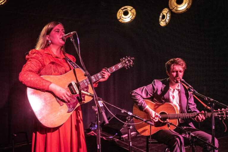 Een vrouw met een gitaar en een man met een gitaar voeren een live muziekoptreden uit op het podium.