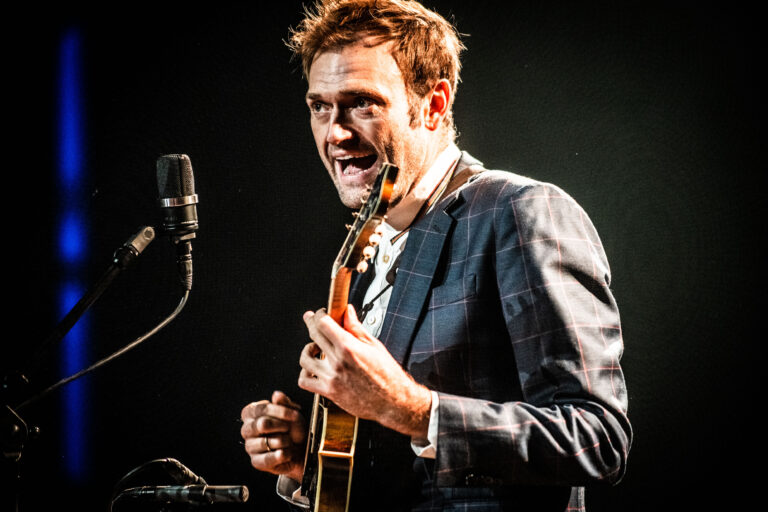 Een man in een donker kostuum zingt en speelt gitaar op een podium met een microfoon.