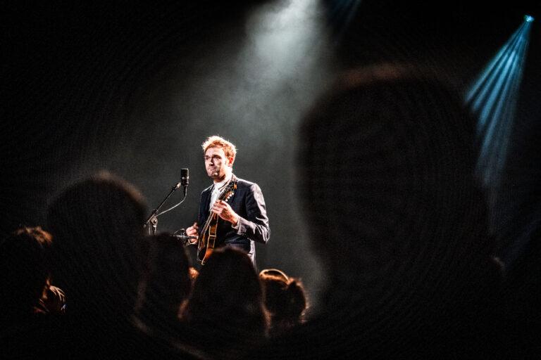 Een muzikant performt op het podium met een gitaar, terwijl het publiek in de schaduw toekijkt.