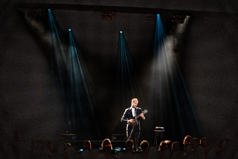 Een solo artiest speelt gitaar op een podium, belicht door gerichte lichtstralen, met een menigte in de zaal.