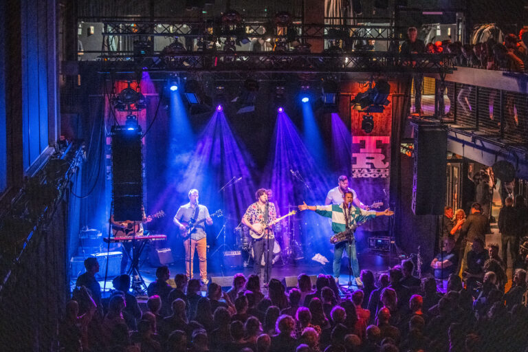 Een band speelt live op een podium met kleurrijke verlichting voor een enthousiast publiek.