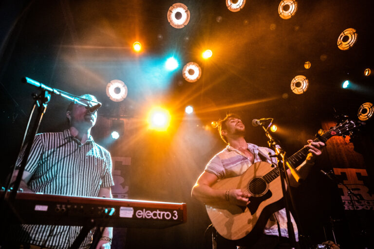 Twee muzikanten optreden op een podium verlicht door kleurrijke lichten, met één persoon die gitaar speelt en de ander die een synthesizer bespeelt.