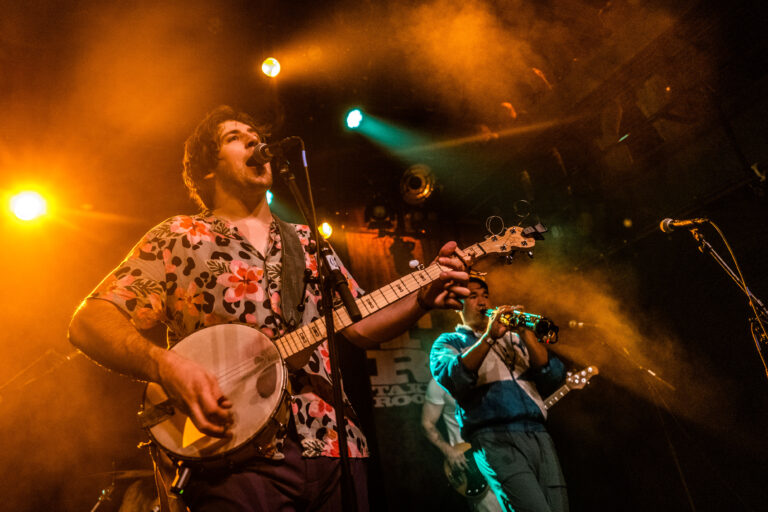 Een muzikant met een banjo zingt op het podium terwijl een andere muzikant op de achtergrond speelt, verlicht door gekleurd licht.