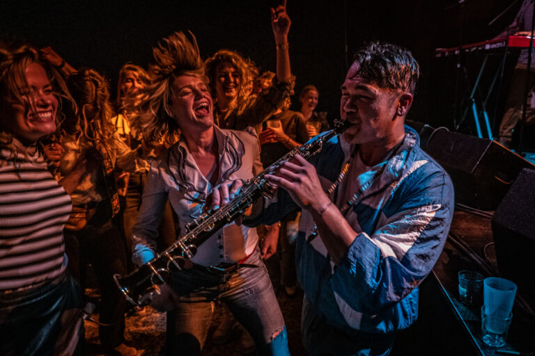 Een muzikant speelt saxofon terwijl de menigte danst en enthousiast is in een sfeervolle omgeving.
