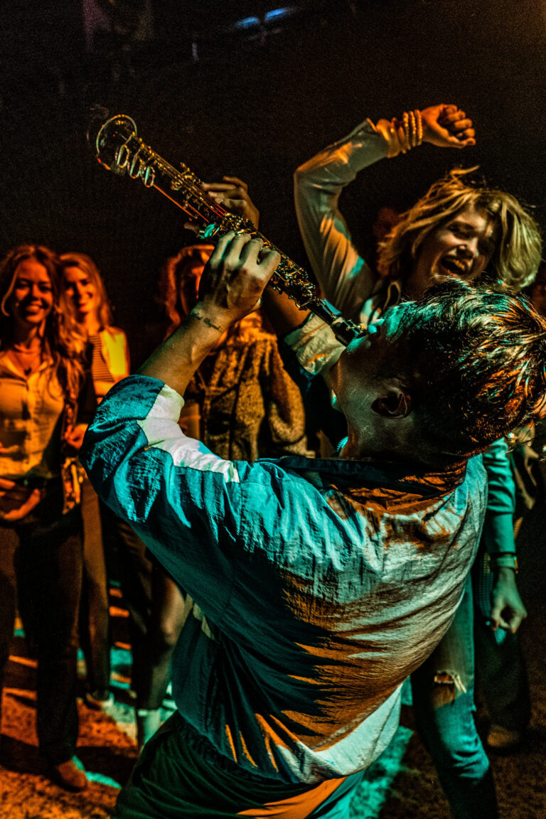 Een saxofonist speelt enthousiast terwijl mensen om hem heen dansen en plezier hebben op een levendig feest.