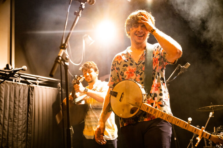 Twee muzikanten op het podium, waarbij de voorste muzikant met een banjo speelt en lacht in het licht van de spotlights.