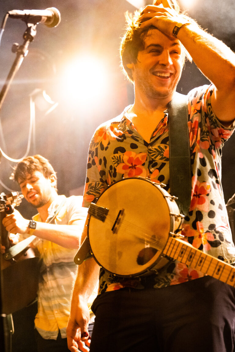 Twee muzikanten op het podium, met een banjo en gitaar, stralend onder de lichten.