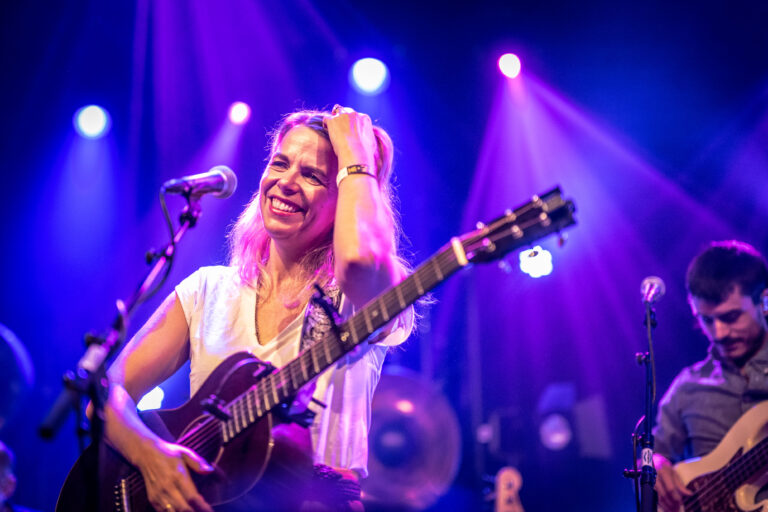 Een blije muzikante met een gitaar staat op het podium, omringd door kleurrijke lichten.