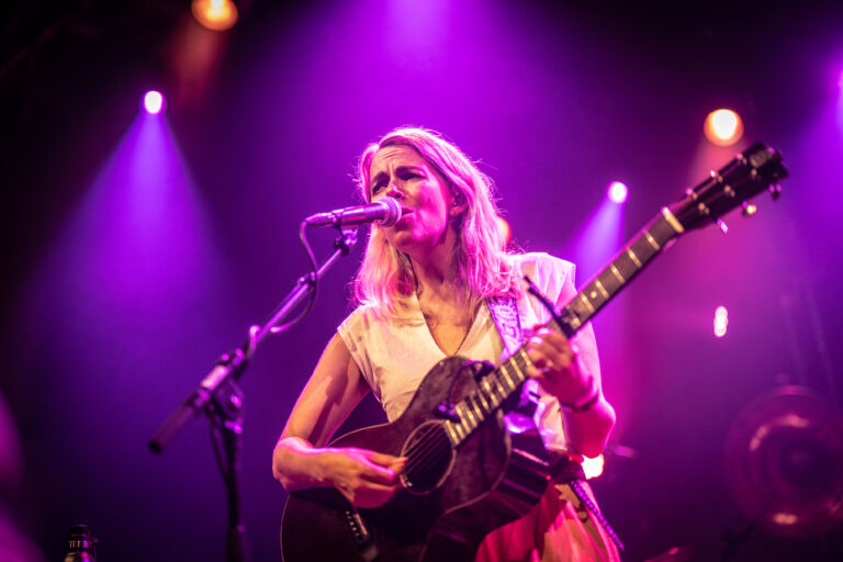 Een vrouwelijke muzikant zingt en speelt gitaar op een podium met kleurrijke verlichting.