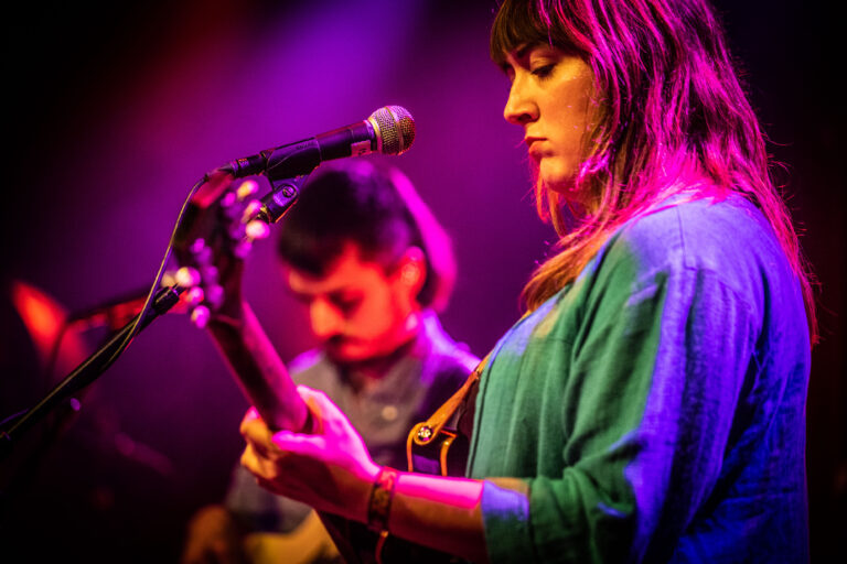 Een muzikant speelt gitaar op het podium, met kleurrijke verlichting op de achtergrond.