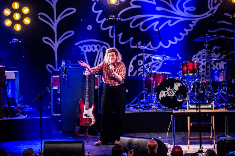 Een performer staat op het podium tijdens een live optreden, met een gitaar en drumstel op de achtergrond.