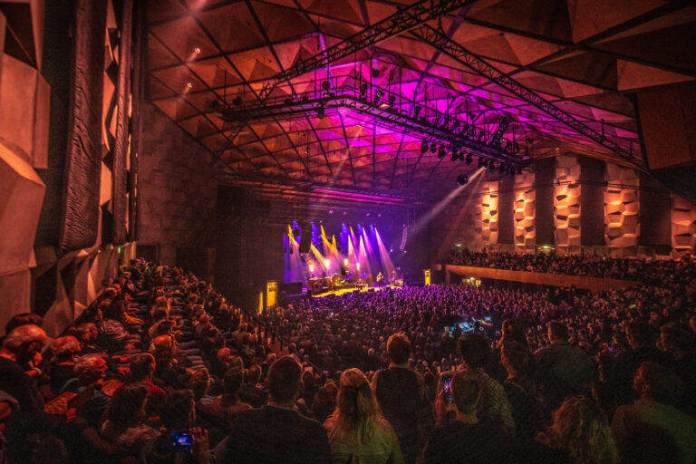 Een druk concert met een publiek dat naar een podium kijkt, verlicht door kleurrijke lichten.