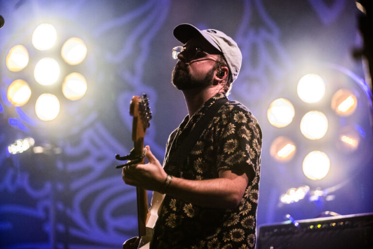 Een muzikant speelt gitaar op een podium met fel licht achter zich.