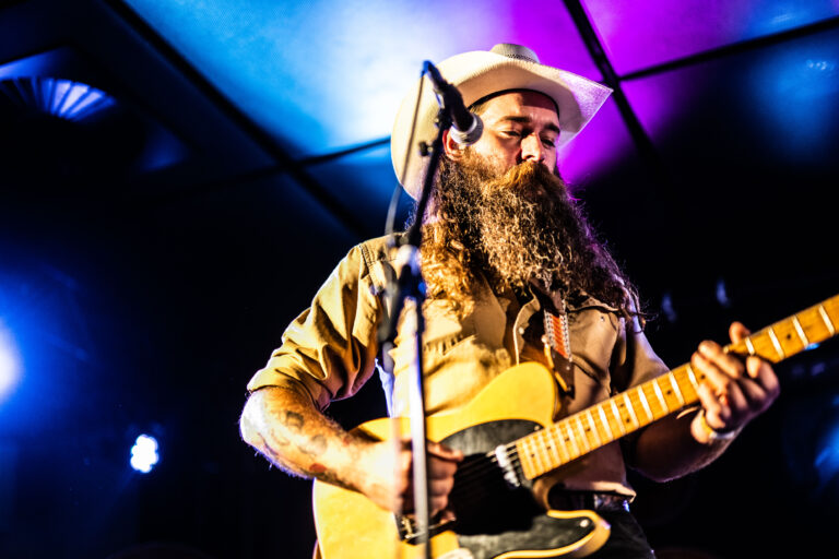 Een man met een lange baard en een hoed speelt gitaar op het podium met kleurrijke verlichting op de achtergrond.