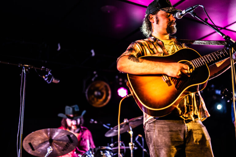 Een muzikant met een gitaar optredend op een podium, met een drummer op de achtergrond.