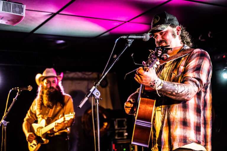 Twee muzikanten optreden op een podium, met één speler die een gitaar bespeelt en zingt terwijl de andere gitaar speelt.