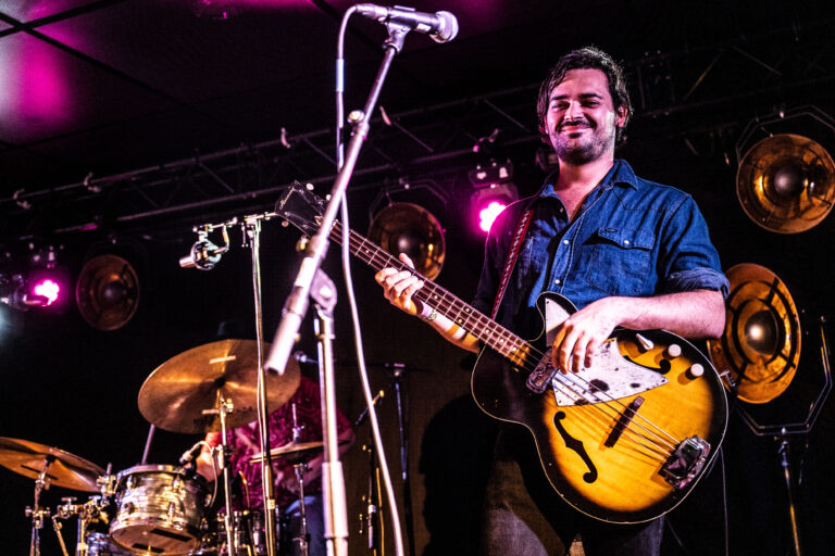 Een muzikant met een basgitaar staat op het podium, terwijl een drummer zichtbaar is op de achtergrond.