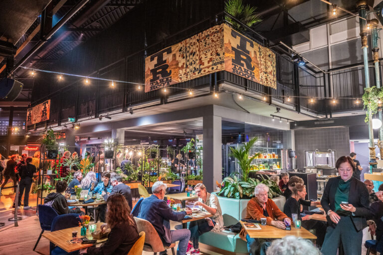 Drukke eetgelegenheid met planten, sfeervolle verlichting en gasten die aan tafels zitten.