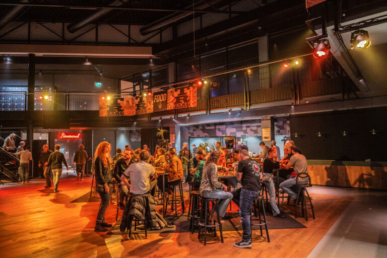 Een drukbezochte horecagelegenheid met bezoekers die aan hoge tafels zitten en met elkaar praten onder sfeervolle verlichting.