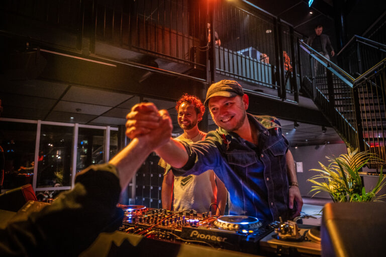 Twee mannen schudden elkaar de hand achter een dj-set in een sfeervolle clubomgeving.