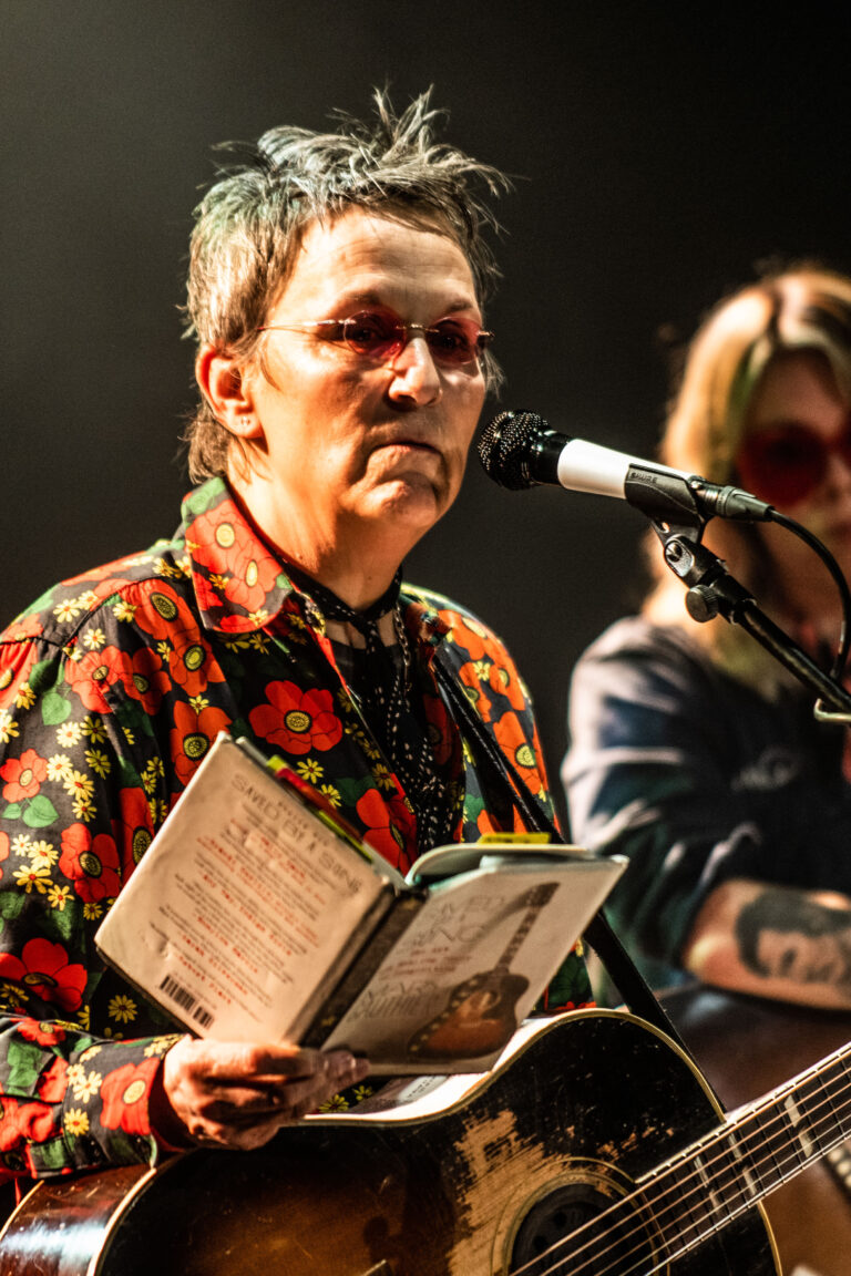 Een muzikant met een bloemenoverhemd die een tekstboek vasthoudt en in een microfoon zingt, terwijl een andere muzikant op de achtergrond speelt.