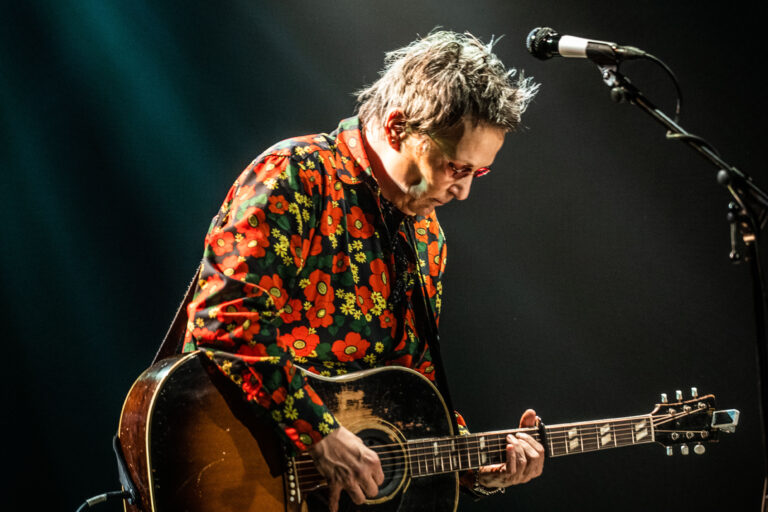 Een muzikant speelt gitaar en draagt een opvallend bloemenpatroon shirt, terwijl hij op het podium staat.