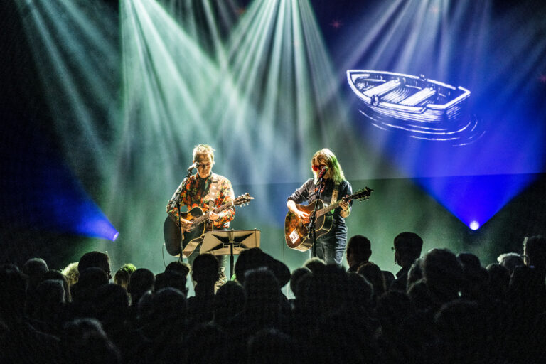 Twee muzikanten met gitaren optreden op een podium verlicht door heldere lichten, met een menigte ervoor.